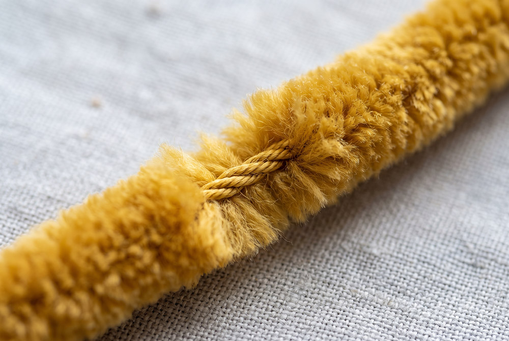 Gros plan sur un fil chenille jaune moutarde : velours dense et âme torsadée visible sur une portion dénudée